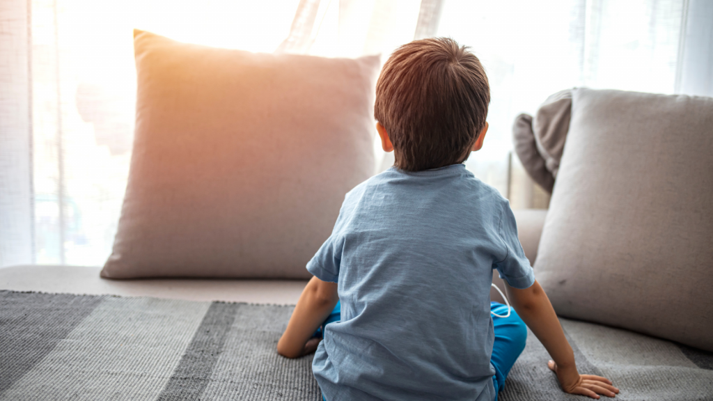 Boy from back, looking trough the window. Little boy sitting on sofa at home