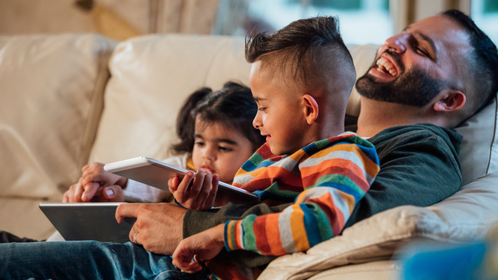 A father and his two children sitting on the couch, watching the screen of a tablet